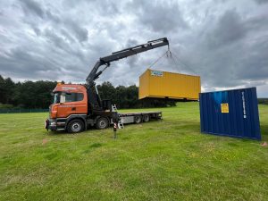a truck lifting up a yellow storage container next to a blue one